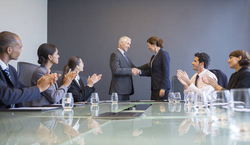 2 people standing shaking hands at a meeting while 4 others are clapping at their seats